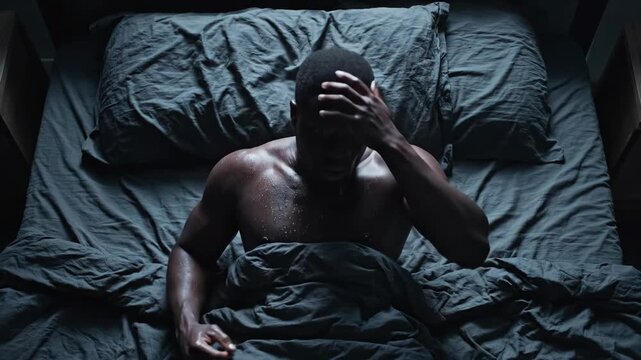 A man wakes up suddenly in a dark room, covered in sweat, looking terrified and disoriented after a vivid nightmare. He clutches the bedsheets, his eyes wide with fear and confusion.