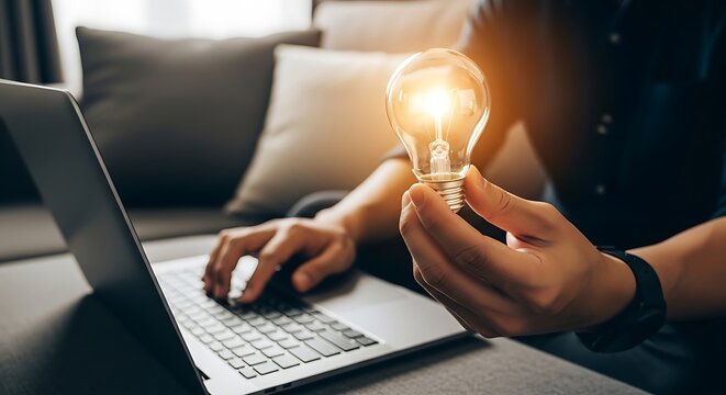 Focused professional holding a glowing light bulb while working on a silver laptop to develop innovative concepts for a new business project that will inspire great future commercial growth.