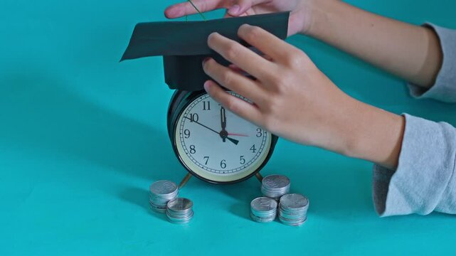 Time to graduate concept featuring alarm clock with academic hat and stacked coins on teal background