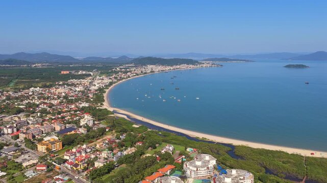 raia da Cachoeira do Bom Jesus in Florianopolis municipality Santa Catarina state South region Brazil with curved bay shoreline urban coastal strip and anchored boats, drone forward glide