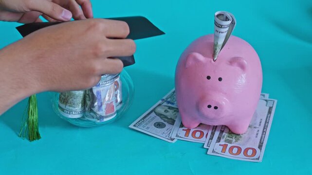Hand placing graduation cap on glass jar filled with cash next to pink piggy bank