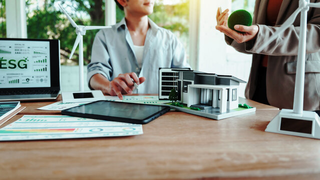 Business meeting on sustainable real estate: ESG dashboard on laptop, model eco house on desk, wind turbines and green concept props in modern office.