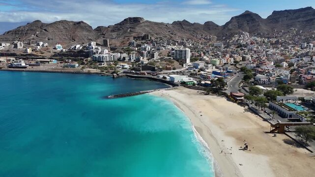 aerial backward drone shot revealing Praia Laginha in Mindelo, Sao Vicente, Cape Verde. Turquoise Atlantic waters, white sand beach, and the coastal city framed by vol