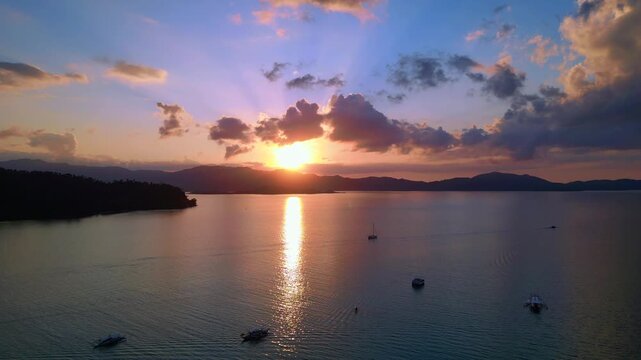 Tropical sun setting behind islands, reflecting golden light on peaceful water with outrigger bangka boats anchored. Lovely aerial view panorama overview drone