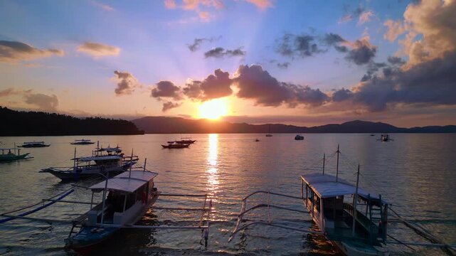 Traditional philipines outrigger bancas resting in a tranquil bay during an impressive colorful sunset, creating a peaceful tropical scene.ascending raise up panorama drone