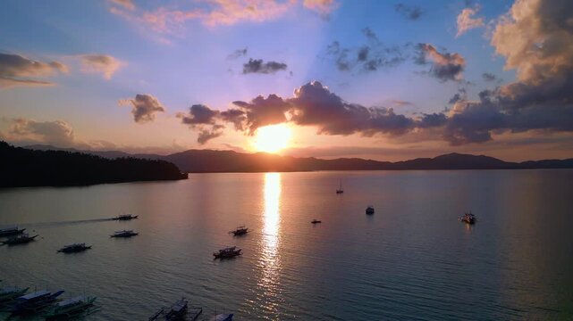 Traditional outrigger bancas resting in a tranquil bay during an impressive colorful sunset, creating a peaceful tropical scene. speed ramp, time lapse. Dramatic aerial view ascending raise up panora