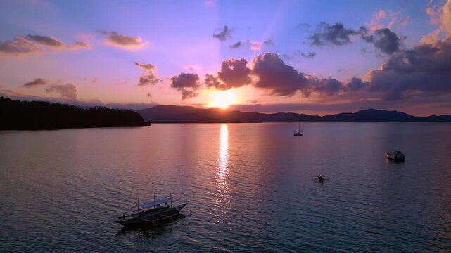 Tropical sun setting behind islands, reflecting golden light on peaceful water with outrigger bangka boats anchored. Great aerial view static panorama hovering drone