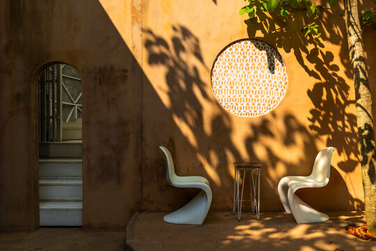 Modern white chairs and a small table set against an orange wall with tree shadows and a circular window in a minimalist outdoor cafe area.