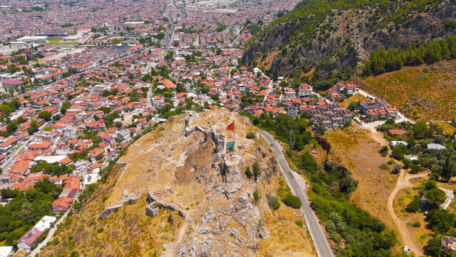 Fethiye, Turkey. Large Turkish flag flying on flagpole atop the historic Fethiye Castle hill with ancient Lycian rock cut tombs in the surrounding cliffs, drone footage.. Aerial View