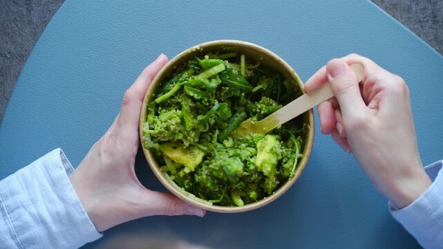 Woman hands mixing fresh vegan salad with quinoa, avocado, pesto dressing in paper bowl, top view, healthy lunch on table representing balanced diet. Mixing green salad with pesto in eco friendly bowl