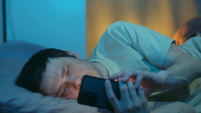 Sleepy man with tired, squinting eyes scrolling on smartphone late at night, lying in bed, fighting sleep while doomscrolling through social media, watching videos. Sleepy man use smartphone, in bed
