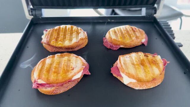 Close up of four delicious grilled panini sandwiches with melted cheese and pastrami meat cooking on a hot griddle with steam rising.