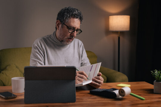 Mature man writing financial notes in planner at home with tablet and calculator