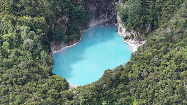 Aerial close view of turquoise crater lake surrounded by dense forest inside Waimangu Volcanic Valley in New Zealand.