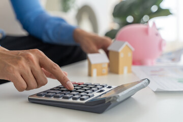 Close up businessman hand using calculator on desk with blurred wooden house models and piggy bank,...