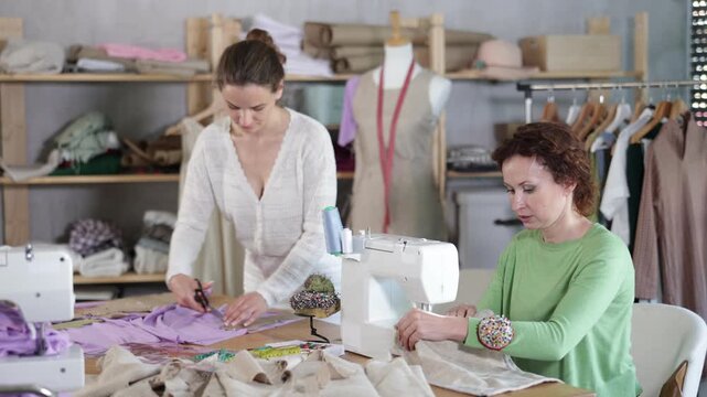 Two skilled female tailors focusing on creating custom garments in small workshop, cutting fabric along paper pattern and assembling pieces with sewing machine