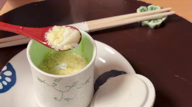 A close up video capturing a red spoon lifting a delicious steamed egg custard chawanmushi from a small ceramic cup.