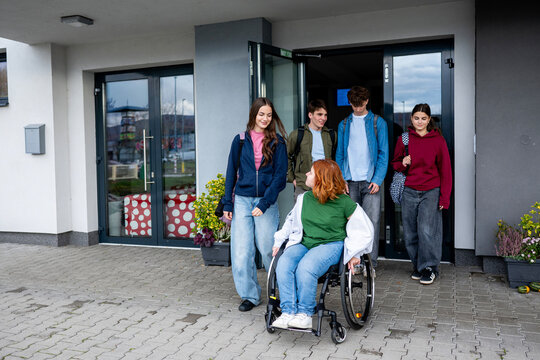 Group of teenage students supporting friend in wheelchair in front of school building.