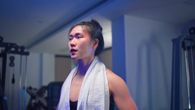 Determined young Asian woman performing lateral raises with dumbbells in a modern gym featuring blue ambient lighting, focusing on fitness and strength training.