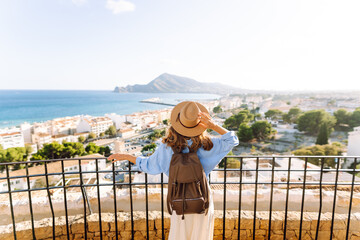 A woman stands by a railing looking at a coastal town. She wears a hat and a backpack. The view shows buildings and mountains in the background under a clear sky. © maxbelchenko