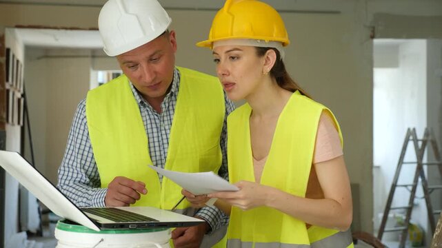 Portrait of woman architect and foreman holding blueprints and using laptop while discussing working process in house being renovated