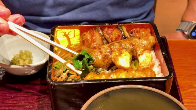Person using chopsticks to eat delicious japanese yakitori bento box with grilled chicken and rice in restaurant.