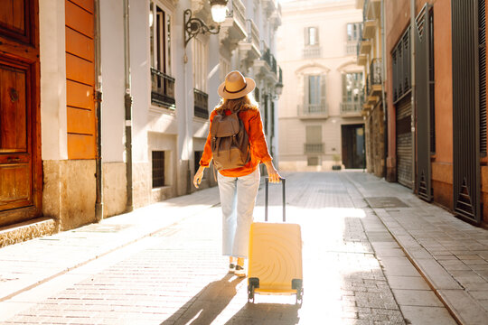 A woman walks along a narrow street, pulling a bright yellow suitcase. She wears a backpack and a wide-brimmed hat. Sunlight shines on the buildings around her.