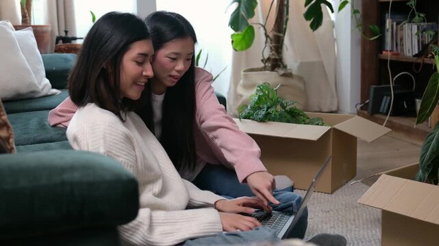 Two cheerful women sitting on the floor with a laptop planning their new apartment