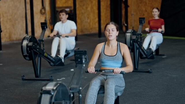 Focused girl works out with rowing machine during class at fitness club. Gym visitor performs exercise, creates load on muscles.