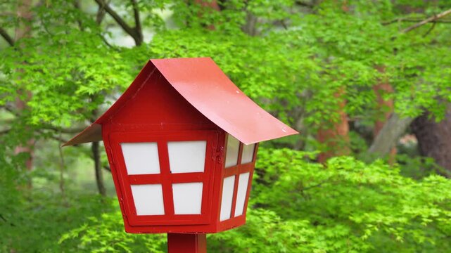 Tiny red-roofed miniature house perched atop a small pillar with lush forest backdrop in Japan, evoking whimsy and serenity