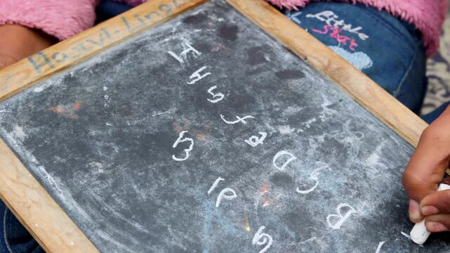 Close-up of an Indian village child writing numbers on a slate board with chalk. A candid rural education moment highlighting early learning, basic numeracy skills, and traditional study methods in a 