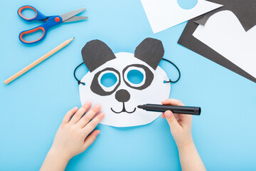 Little child boy hand drawing white black panda bear paper mask with marker on light blue table background. Pastel color. Point of view shot. Closeup. Creating decoration. Top down view. © fotoduets