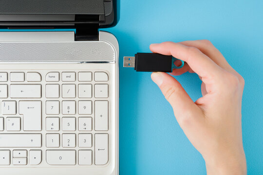 Young adult woman hand fingers holding and putting dark black flash drive into white laptop on light blue table background. Pastel color. Top down view.