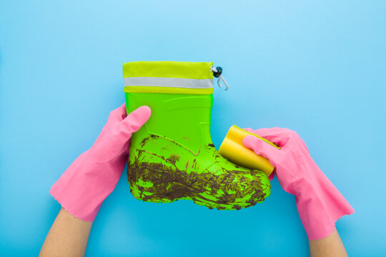Woman hands in pink rubber protective gloves holding sponge and cleaning dirty green kid rubber boot on light blue table background. Pastel color. Closeup. Point of view shot. Flat lay. Top down view.