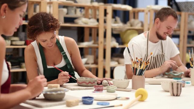 Three pottery masters work together on ceramic tableware, enjoying creative process as they paint mugs in the workshop 