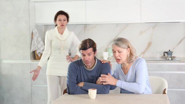 Three people are in bright kitchen, woman expresses her displeasure to man and old woman calms him down