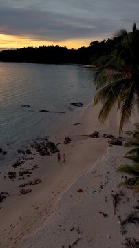 As the sun sets on Koh Kood Island, vibrant colors fill the sky while a couple strolls along the pristine beach. Gentle waves and swaying palm trees create a serene atmosphere.