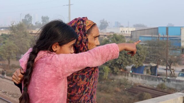 A young Indian girl points into the distance while standing beside her grandmother, capturing a meaningful intergenerational moment of curiosity and guidance in a rural village setting