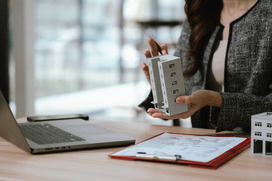 Businesswoman holding a small building model at a desk with a laptop and documents, representing real estate investment, property management, and financial decision-making.