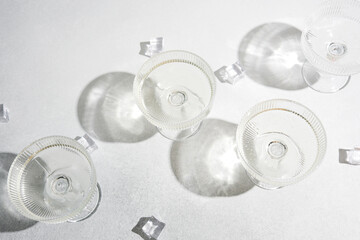 Obraz premium Four coupe glasses with clear liquid and ice cubes on a textured white surface creating strong shadows.