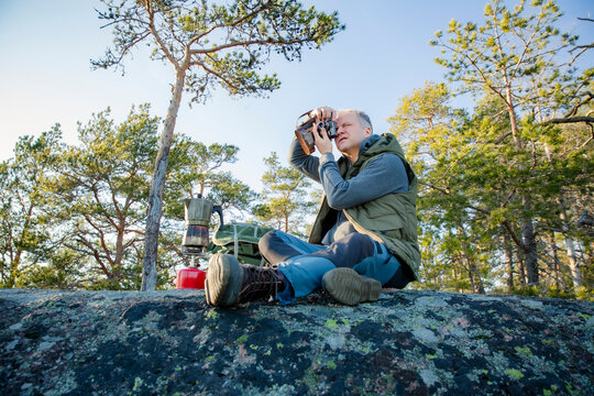 Male hiker taking photos with vintage camera on scenic cliff in Finland. Travel photography, adventure tourism, outdoor lifestyle