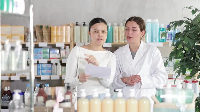 Thoughtful Asian woman in pharmacy holding printed doctor prescription and asking polite female pharmacist for assistance in finding listed medications