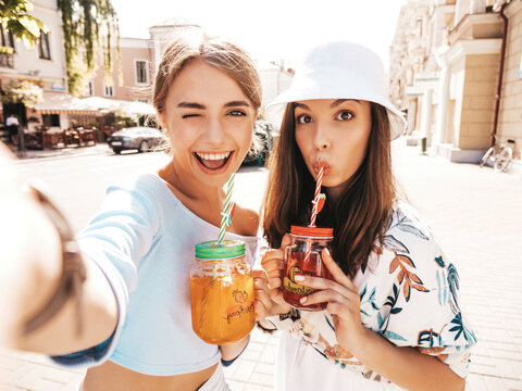 Two young beautiful smiling hipster in trendy summer clothes.Panama hat.Sexy carefree women taking selfie portrait photos on smartphone in the street.Drinking fresh cocktail smoozy drink