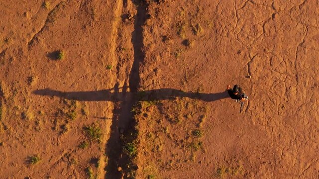 Drone top down view of person walking across red dirt desert landscape casting long dramatic shadow. Golden hour sunlight illuminates barren terrain with sparse vegetation patches.
