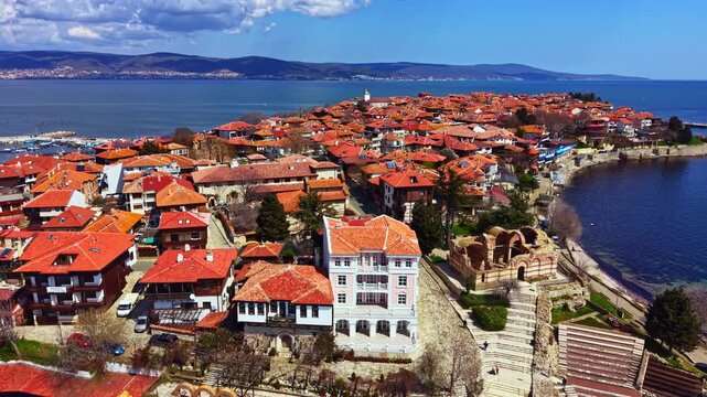 People walk along the coastal path in Nesebar Old Town. The scene shows red-roofed buildings and the sea in bright sunlight. Boats are visible in the harbor, adding to the lively atmosphere.