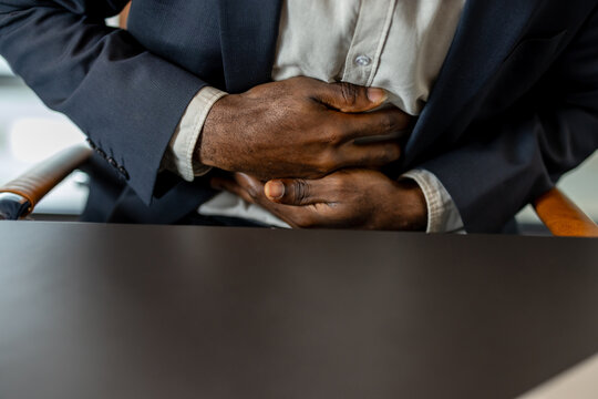 Businessman in suit seated at desk clutching his abdomen in visible discomfort, depicting indigestion, heartburn, constipation and work-related digestive distress and stress symptoms