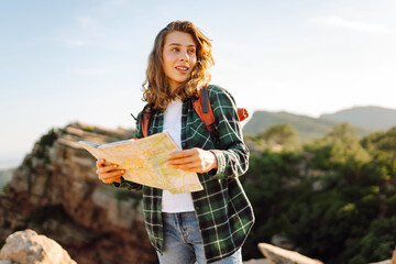 A hiker with a backpack stands on a mountain with a map in her hands. A young woman with a map looks for directions while hiking. Concepts of enjoyment, navigation. Active lifestyle. © maxbelchenko
