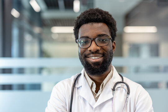 Male professional doctor looking at camera and smiling, wearing a white lab coat and stethoscope, posing against a modern clinic background, representing healthcare expertise and patient care