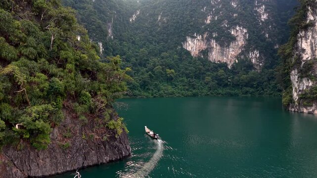 Experience enchanting views of Khao Sok National Park in Thailand. A boat glides smoothly through emerald waters surrounded by towering limestone cliffs and rich greenery.
