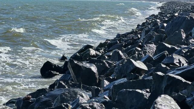 Waves hit dark rocks along the shoreline while the sun shines. Soft sounds of water mingle with the changing tides. A clear view of the coast is visible in the background.
